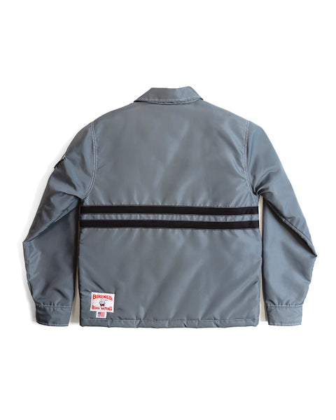 Back of Quilt Lined Comp Jacket in Slate with two horizontal black stripes across the chest with contrast white stitching on white background. Bottom left of the jacket with Birdwell License Plate Label with smaller made in USA label beneath.