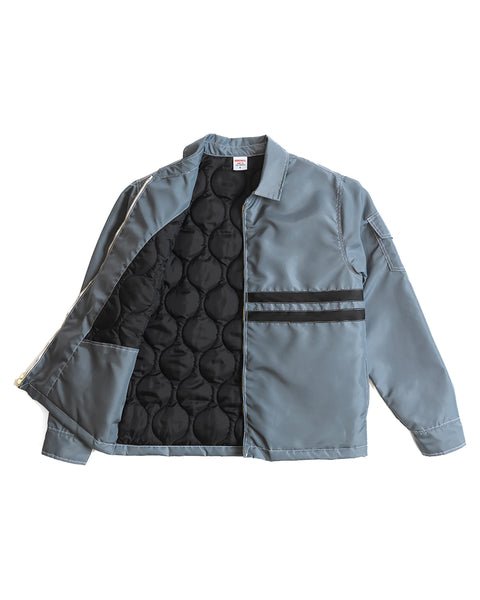 Front of Quilt Lined Comp Jacket in Slate with two horizontal black stripes across the chest with contrast white stitching on white background. Left side of the jacket open to show the insulated interior.