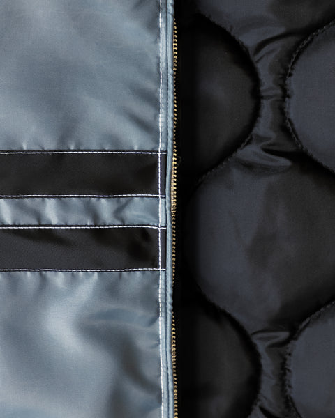 Close up detail shot of the material of the jacket. Left of the image shows two horizontal black stripes with white contrast stitching on the slate jacket. Center of the image shows the gold talon zipper and right side of the image shows the black insulated interior of the jacket.