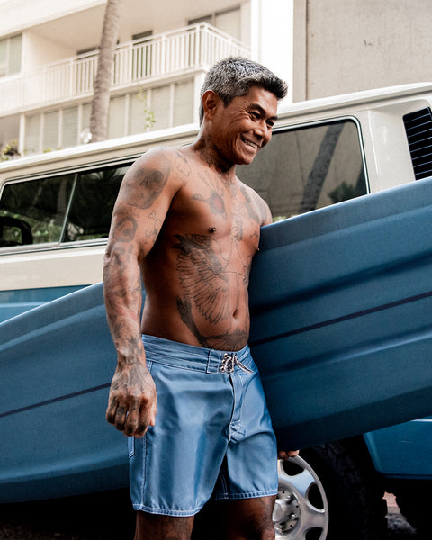 Toots wearing the 310 Boardshort in Federal Blue while holding a blue surfboard while standing in front of a two tone vw bus with white top and blue bottom.