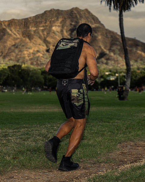 Bill Maeda running in the grass with mountain and palm trees in the background wearing the GR1 backpack and 300 Boardshorts in Black and Camo.