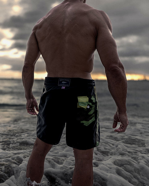 Bill Maeda standing in the ocean with dark grey clouds and sky while wearing 300 Boardshorts in Black and Camo with his back facing the viewer while looking out into the ocean.