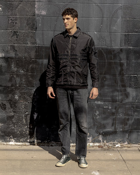 Model wearing the Comp Stripe 25 Jacket Black on Black. Model facing the camera with hands to the side with the front of jacket with pocket on right. Model is wearing black jeans and black converse shoes. Background is brick wall painted black.