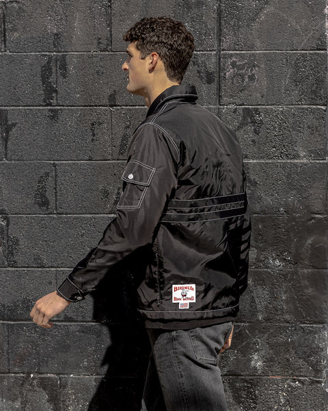 Model wearing the Comp Stripe 25 Jacket Black on Black. Model walking to the left with a side view of the jacket with pocket and license plate label facing the camera. Background is brick wall painted black.