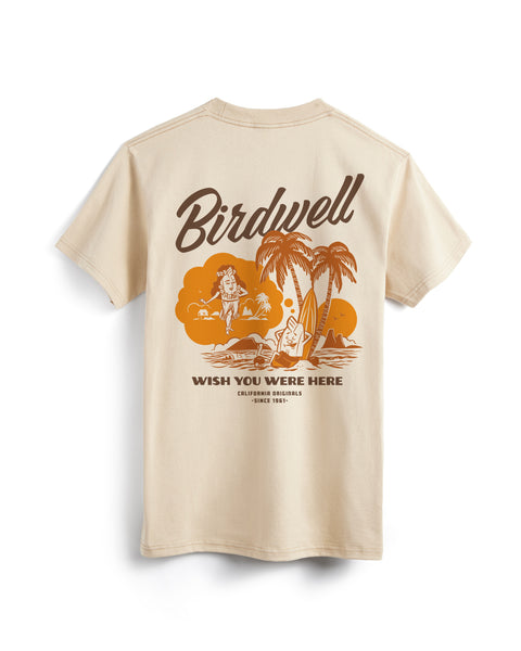 Back view of sand colored t-shirt on white background. Screen-printed illustration of Birdie leaning up against a palm tree and surfboard while dreaming of a female birdie in a hula skirt. Birdwell in script font above the trees and 