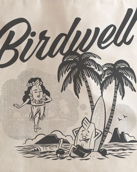 Close up detail shot of the Screen Printed Daydreamin Birdie Graphic in Grey with Birdwell in Script Text and Birdie with his eyes closed leaning up against a Surfboard and two palm trees on the beach while dreaming of a female Birdie in a hula skirt. Background is bone colored canvas.