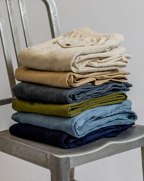 Stack of pants on a silver metal chair in front of white background. Top of stack is sand balboa pants followed by tan coronado pant, slate balboa pant, army green coronado pant, blue balboa pant and dark blue balboa pant.