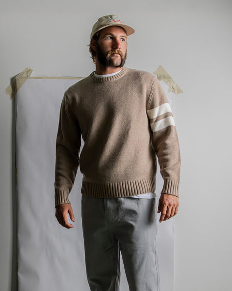 Model wearing oatmeal colored sweater with two horizontal cream stripes over white t-shirt and quarry pants and tan hat in front of white wall with white paper taped to wall.