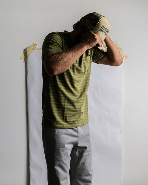 Model wearing light green hat with camo brim with Birdwell Beach Britches logo screenprinted on front of hat with green pocket shirt with thin horizontal yellow and green stripes and quarry pants in front of wall with white paper taped to white wall.