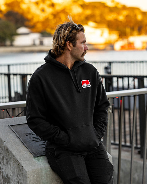 Kevin looking to the right wearing the Grit Hoodie in Black and black pants with hands in pockets leaning against a cement block. Harbor in background with trees and golden sun cast upon the trees.