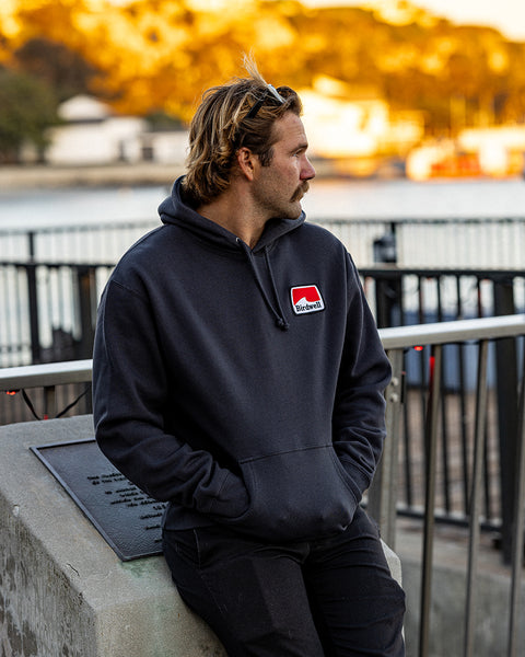 Kevin looking to the right wearing the Grit Hoodie and black pants with hands in pockets leaning against a cement block. Harbor in background with trees and golden sun cast upon the trees.