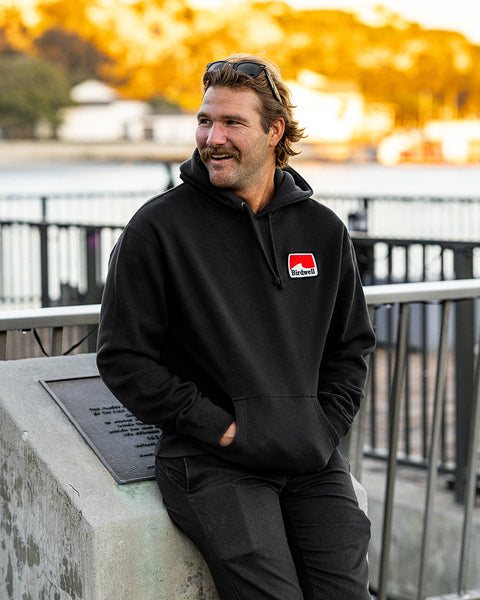 Kevin looking to the left wearing the Grit Hoodie in Black and black pants with hands in pockets leaning against a cement block. Harbor in background with trees and golden sun cast upon the trees.