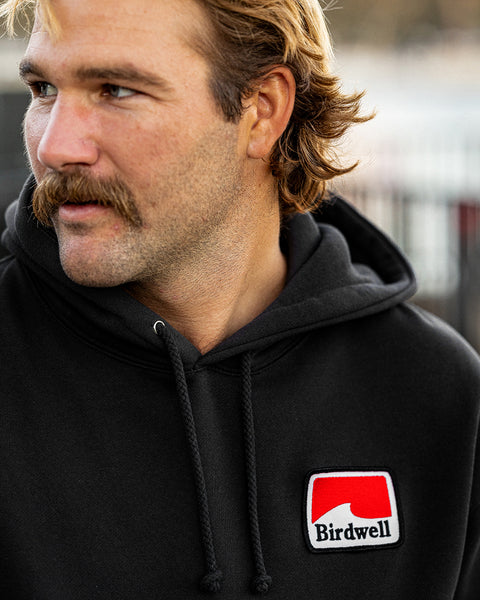 Close up of Kevin looking to the left wearing the Grit Hoodie with patch in lower right hand corner. Rectangular patch has geometric white wave with red background and Birdwell embroidered in black.