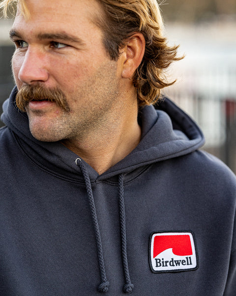 Close up of Kevin looking to the left wearing the Grit Hoodie with patch in lower right hand corner. Rectangular patch has geometric white wave with red background and Birdwell embroidered in black.