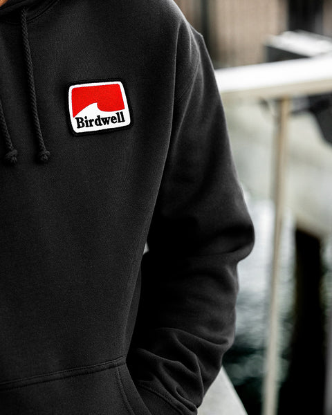 Close up of he Grit Hoodie with patch in the left chest area. Rectangular patch has geometric white wave with red background and Birdwell embroidered in black.