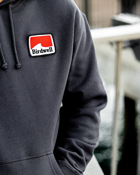 Close up of he Grit Hoodie with patch in the left chest area. Rectangular patch has geometric white wave with red background and Birdwell embroidered in black.