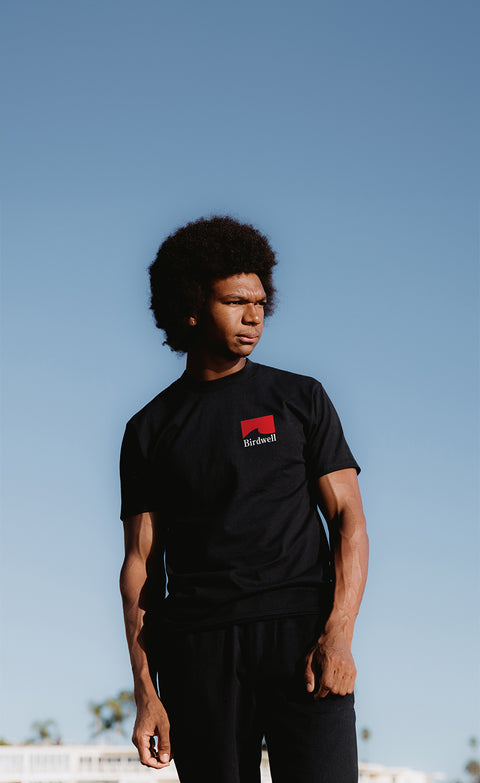 Obi wearing the grit t-shirt in black. T-Shirt has a red wave with white birdwell text. Blue sky in background with white homes and palm trees towards the bottom of the image.