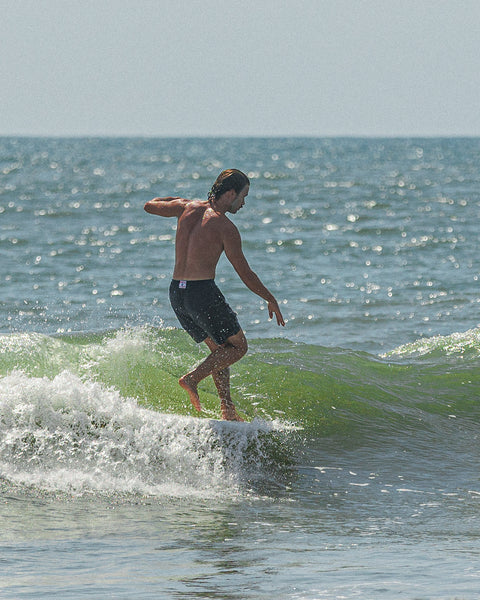 Surfer wearing the black Wright Shorts, surfer is angled to the right at the tip of the board. Background showing blue-green waves and blue sky.