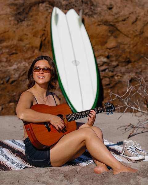 Woman wearing the Birdie Boardshorts with 4-way stretch on a towel sitting in the sand at the beach. She is also wearing sunglasses and holding a guitar with rocks and surfboard in the background.