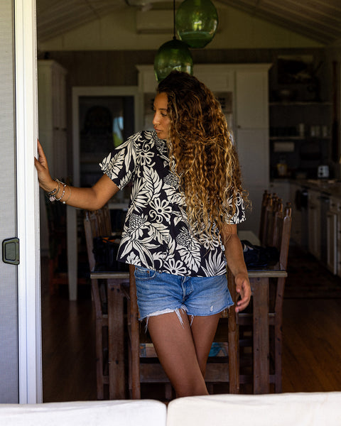 Woman is wearing a black and white floral printed button up woven top with denim shorts while standing in a door frame. In the background there is a dining room and kitchen with two green pendant lights hanging from the ceiling.