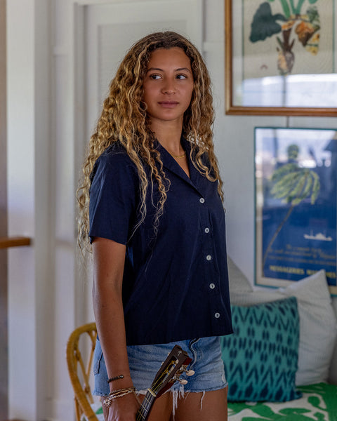 Woman is wearing a navy button up shirt and denim shorts in a house with framed pictures of plants in the background and a chair with pillows on it. The pillows are blue, green, and white. She is also holding a guitar that is peaking through the bottom of the image.