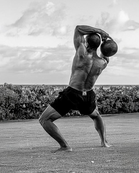 Bill Maeda wearing the Wright Lined Short in Black. Bill is lifting a kettlebell weight over his shoulders. Image is black and white. Background is grass with a row of bushes and clouds in the sky.