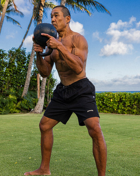 Front view of Bill Maeda wearing the Wright Lined Short in Black. Bill is lifting a kettlebell weight at his chest. Background is green grass with a row of bushes, palm trees on the left, and clouds in the blue sky.